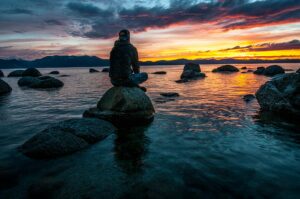 person sitting on rock by water at sunset with colorful sky