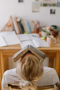 Blonde woman reclining with a book covering her face at a desk.