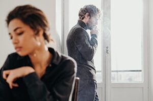 Woman sitting pensively in foreground with man smoking by window