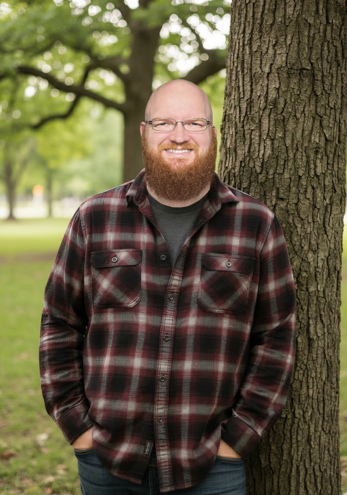 Bearded man with glasses in plaid shirt standing by tree in park.