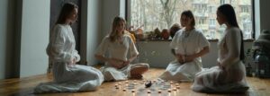 Four women in white dresses meditate with candles on a wooden floor indoors.