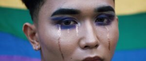 Close-up of a person with makeup crying against a rainbow flag backdrop.